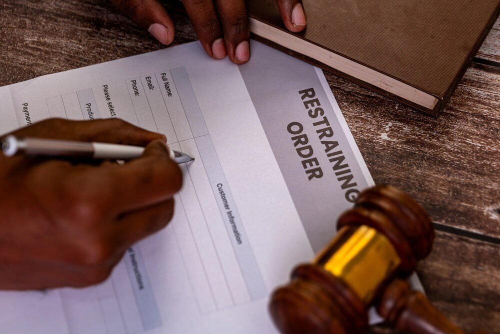 A person filling out a restraining order form with a pen. The form is on a wooden table beside a wooden gavel and a closed book. The person's hand and part of their arm are visible.