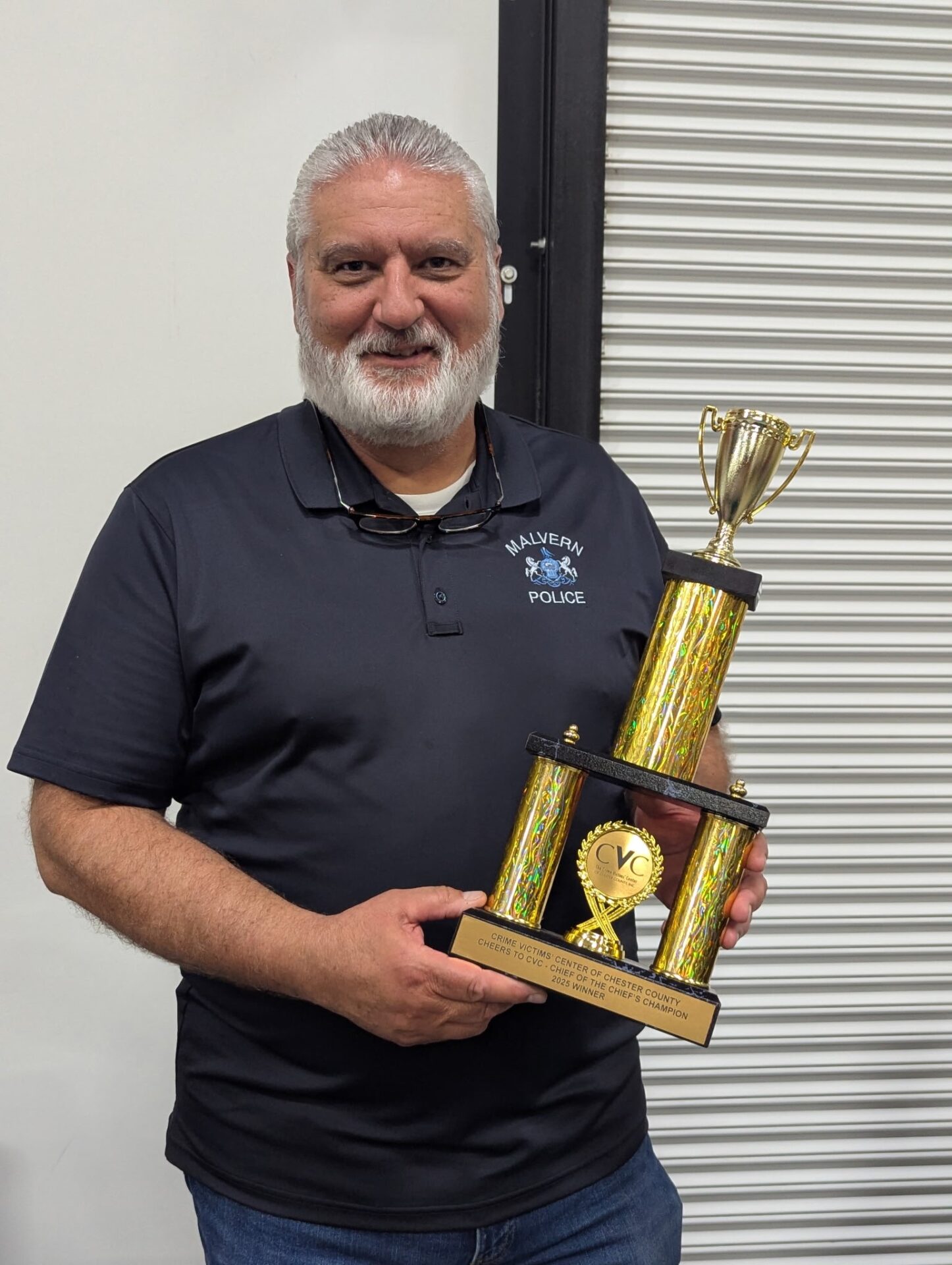 A man with gray hair and a beard wearing a navy blue "Malvern Police" polo shirt smiles while holding a large gold and yellow trophy in front of a white metal rolling door.