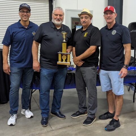 Four men stand together indoors, smiling at the camera. One man in the center holds a trophy. They wear casual clothes and hats, and a table with a blue cloth is behind them. A closed garage door is in the background.
