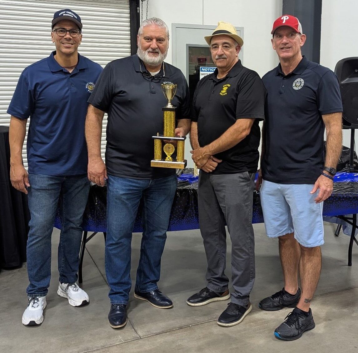 Four men stand together indoors, smiling at the camera. One man in the center holds a trophy. They wear casual clothes and hats, and a table with a blue cloth is behind them. A closed garage door is in the background.