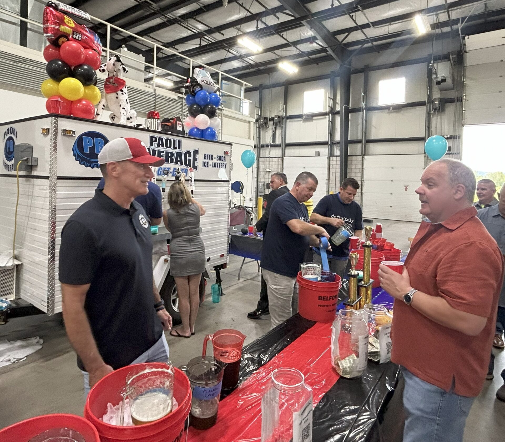 Two men converse at a drink stand inside a large garage, with beverage containers, tip jars, and party decorations including balloons and inflatable cows visible on and around a beverage truck in the background.