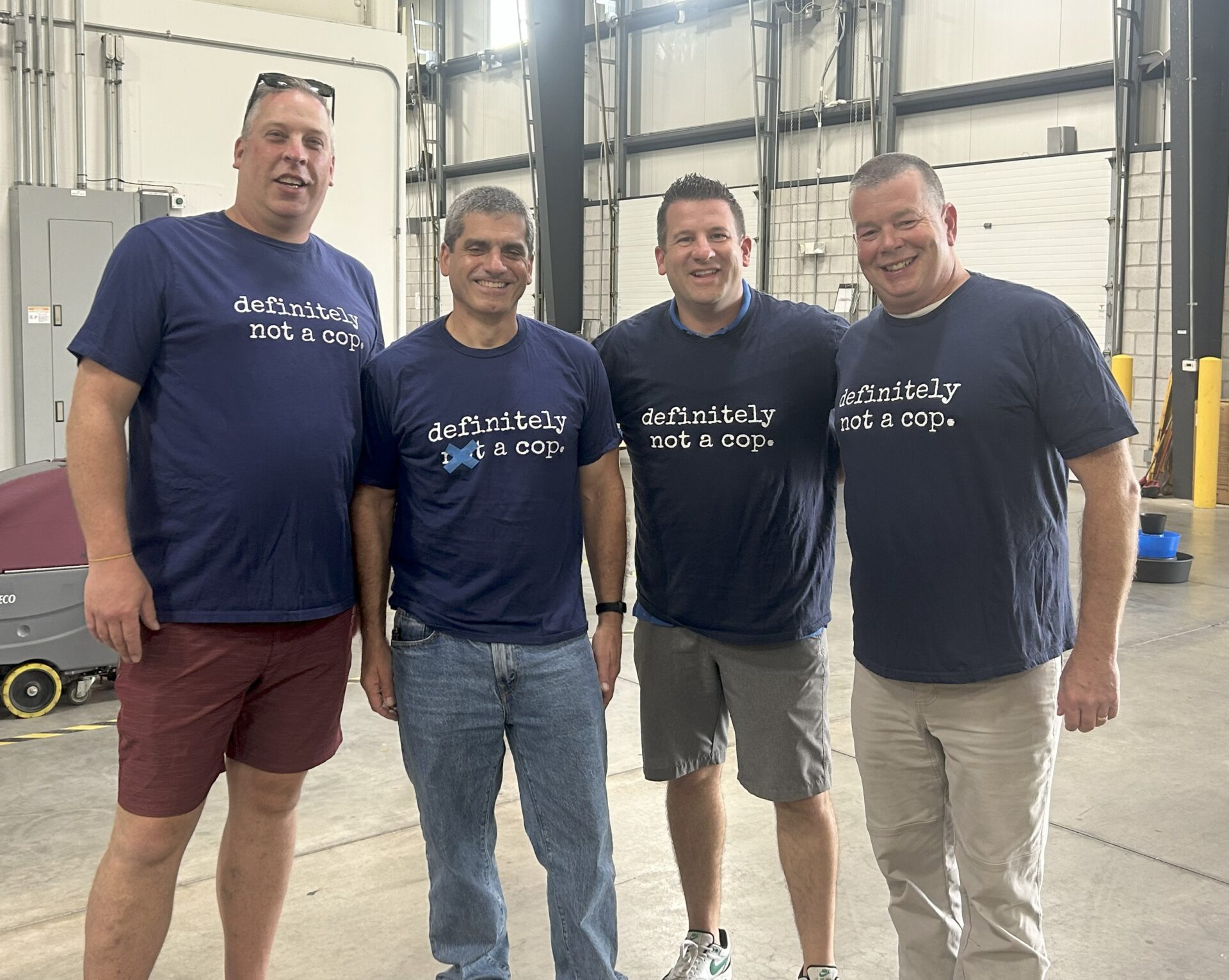 Four men standing indoors, smiling, all wearing matching navy blue shirts that say "definitely not a cop." The setting appears to be an industrial or warehouse space with concrete floors.