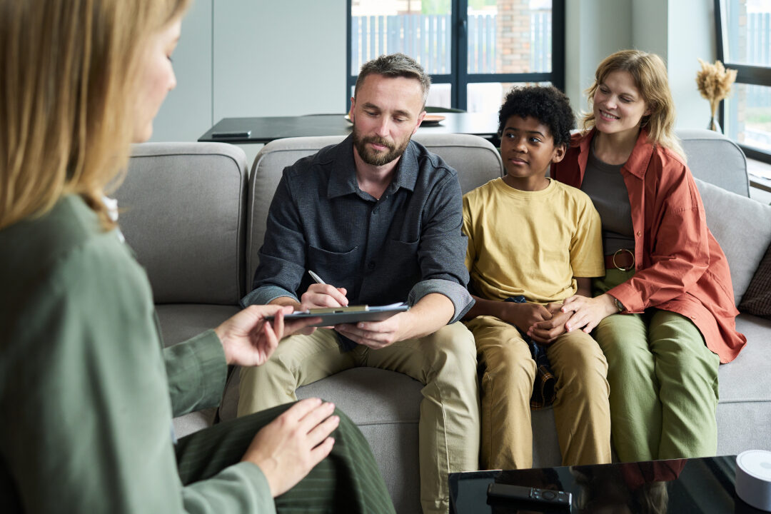 A family of three, sitting on a sofa, talks to a professional holding a clipboard. The father is writing, the child sits between parents, and the mother smiles. The room is modern with a coffee table and large windows.