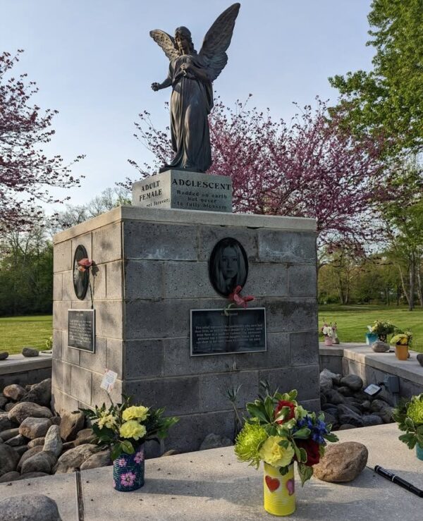 A statue of an angel atop a stone pedestal, surrounded by flowers in vases. Plaques and a carved female portrait adorn the pedestal. Trees with pink blossoms are in the background under a clear sky.