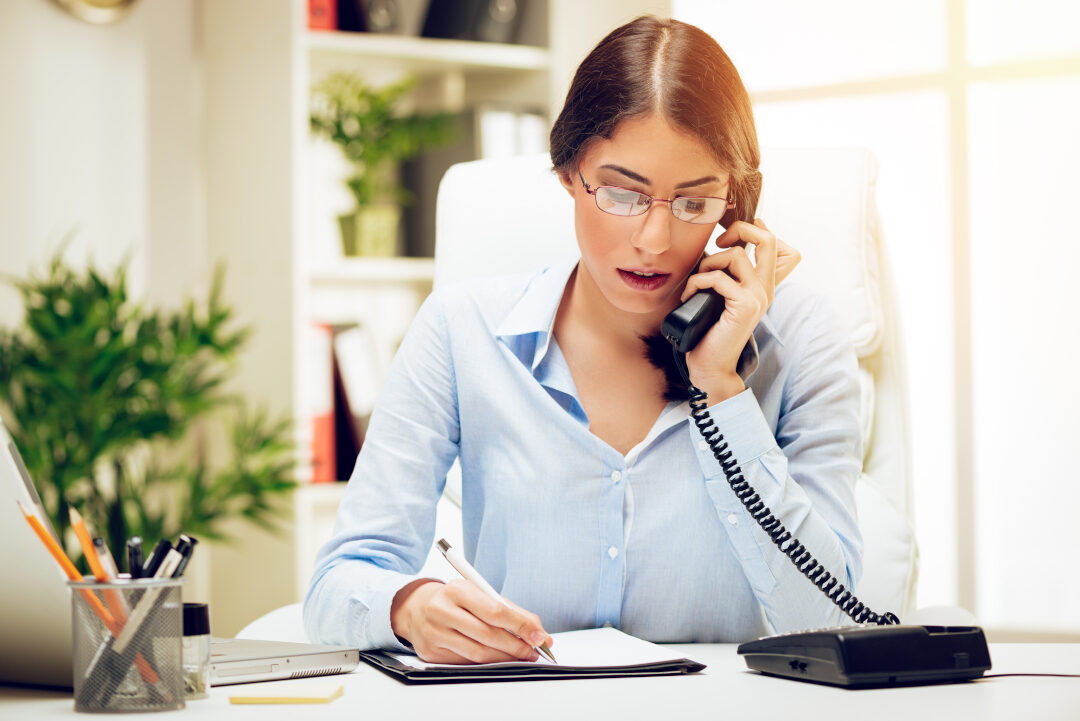 A woman in a blue shirt is sitting at a desk in an office, talking on a landline phone while taking notes on a clipboard. She is surrounded by office supplies, and there is a potted plant in the background.